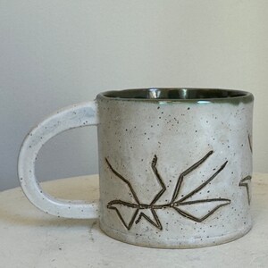 May include: White ceramic mug with a green rim and a brown stick insect design.