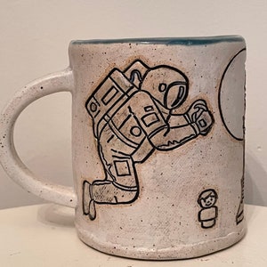 May include: White ceramic mug with a black and white illustration of an astronaut kneeling on the moon. The astronaut is wearing a space suit and has a backpack. There is a small figure of a person standing on the moon.