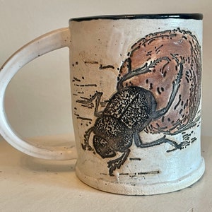May include: White ceramic mug with a black rim and handle. The mug features a brown and black illustration of a beetle crawling on a brown rock.