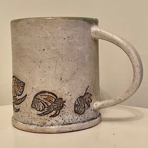 May include: A white ceramic mug with a speckled glaze and a brown handle. The mug has a design of three hermit crabs on the side.
