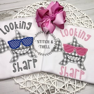May include: Two white t-shirts with a gray and white checkered shark design. One shark has blue sunglasses and the other has pink sunglasses. The text "Looking Sharp" is embroidered on each shirt.