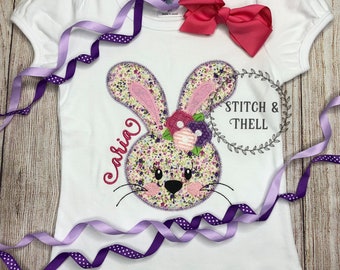 May include: White t-shirt with a pink and purple embroidered bunny face, a floral headband, and the name "Olivia" stitched in purple. The text "Stitch & Thell" is also embroidered on the shirt.