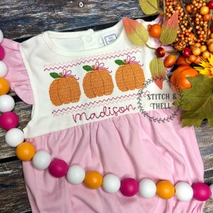 May include: A pink baby onesie with white ruffle sleeves and a white bodice featuring an embroidered design of three orange pumpkins with green stems and a pink bow. The onesie has the name "madison" embroidered below the pumpkins.