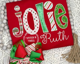 May include: Red Christmas-themed shirt with the name "Jolie Ruth" embroidered in white. The letters are decorated with green, red, and white candy cane and polka dot patterns.