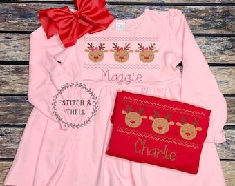 May include: Two pink and red Christmas dresses with reindeer embroidery. The pink dress has the name "Maggie" embroidered on it. The red dress has the name "Charlie" embroidered on it.