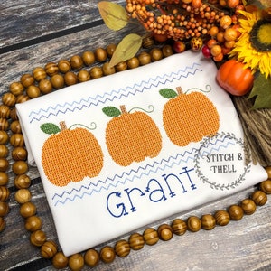 May include: White onesie with three embroidered orange pumpkins and the name "Grant" in blue embroidery.