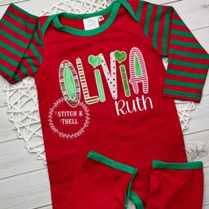 May include: Red and green striped long-sleeve baby onesie with the name "Olivia Ruth" embroidered on the front. The onesie has a green trim around the neckline and the sleeves. The letters are decorated with a Christmas theme.
