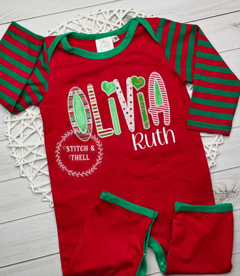 May include: Red and green striped long-sleeve baby onesie with the name "Olivia Ruth" embroidered on the front. The onesie has a green trim around the neckline and the sleeves. The letters are decorated with a Christmas theme.