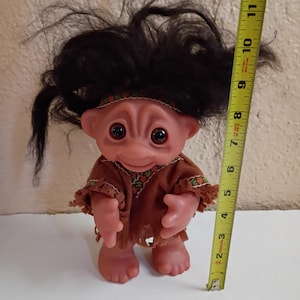 Puede incluir: Una mu&ntilde;eca troll vintage con cabello casta&ntilde;o y un atuendo marr&oacute;n. La mu&ntilde;eca tiene ojos grandes y redondos y mide aproximadamente 15 cm de alto, medidos con una cinta m&eacute;trica amarilla.