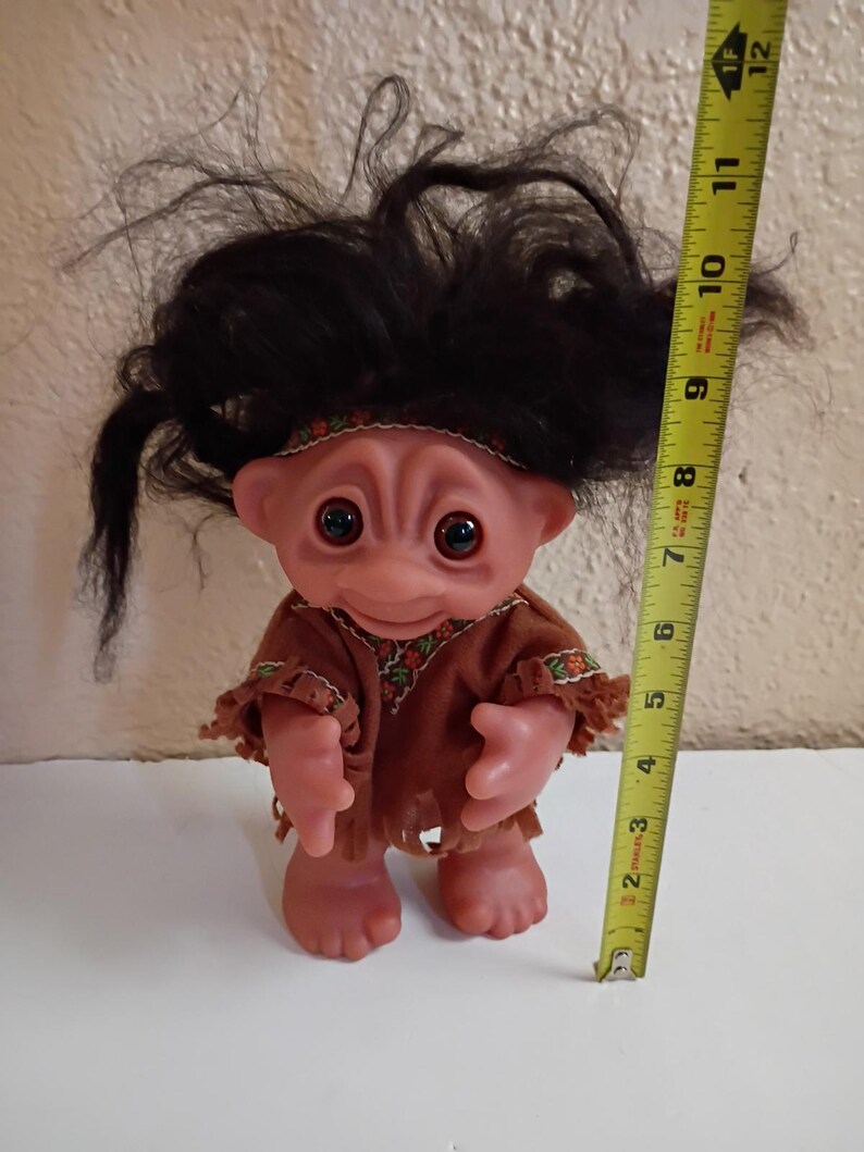 Puede incluir: Una mu&ntilde;eca troll vintage con cabello casta&ntilde;o y un atuendo marr&oacute;n. La mu&ntilde;eca tiene ojos grandes y redondos y mide aproximadamente 15 cm de alto, medidos con una cinta m&eacute;trica amarilla.