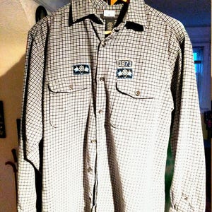May include: A long-sleeved plaid flannel shirt with a black and white checkered pattern. The shirt has two embroidered patches on the front, one with the text "1971" and the other with a graphic design.