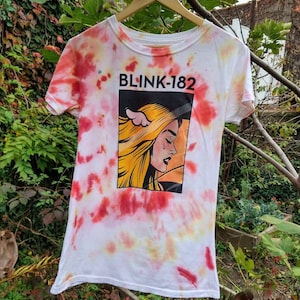 May include: White tie-dye t-shirt with a red, yellow, and black graphic print of a woman with a tear on her cheek. The graphic includes the text "BLINK-182".