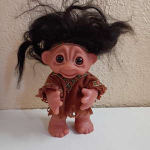 Puede incluir: Una mu&ntilde;eca troll vintage con tez rosada, ojos grandes y una expresi&oacute;n alegre. La mu&ntilde;eca tiene un atuendo marr&oacute;n con flecos y una diadema. Su cabello negro est&aacute; peinado de forma salvaje y puntiaguda. La mu&ntilde;eca est&aacute; de pie sobre una superficie blanca.