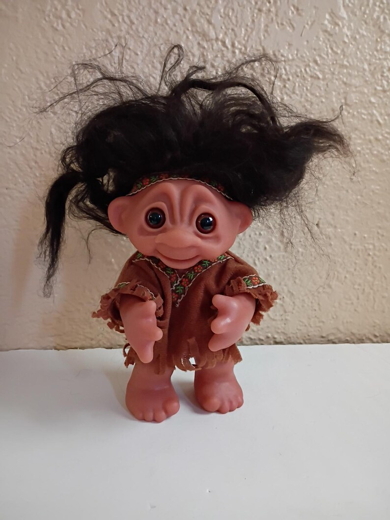 Puede incluir: Una mu&ntilde;eca troll vintage con tez rosada, ojos grandes y una expresi&oacute;n alegre. La mu&ntilde;eca tiene un atuendo marr&oacute;n con flecos y una diadema. Su cabello negro est&aacute; peinado de forma salvaje y puntiaguda. La mu&ntilde;eca est&aacute; de pie sobre una superficie blanca.