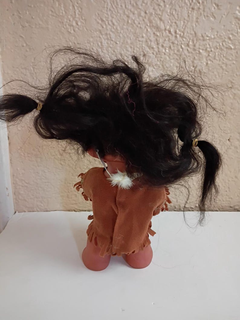 Puede incluir: Una mu&ntilde;eca vintage con cabello oscuro peinado en dos coletas. La mu&ntilde;eca lleva un chaleco marr&oacute;n tipo ante con flecos y un collar de plumas blancas. La piel de la mu&ntilde;eca es de color melocot&oacute;n claro.