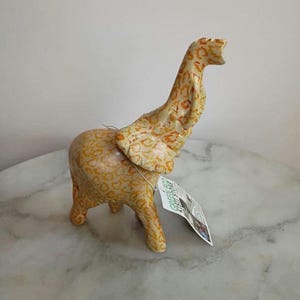 May include: A ceramic giraffe figurine with a cream and orange spotted pattern. The giraffe has a long neck and is standing on a marble surface. A tag is attached to the figurine.
