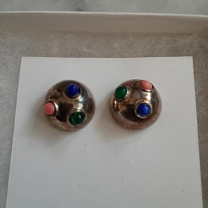 May include: A pair of round, metallic dome-shaped earrings featuring a hammered texture. Each earring is accented with three small, smooth, cabochon-style stones in vibrant shades of blue, green, and pink, set securely into the polished metal surface.