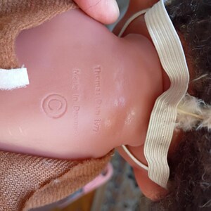 Puede incluir: Parte trasera de una mu&ntilde;eca vintage con el texto "Thomas Dam 1977" y "Made in Denmark". La mu&ntilde;eca tiene un cuerpo de pl&aacute;stico rosa, ropa de tela marr&oacute;n y una correa el&aacute;stica blanca. El pelo de la mu&ntilde;eca es oscuro con plumas blancas.