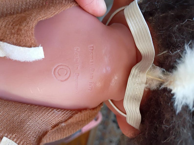 Puede incluir: Parte trasera de una mu&ntilde;eca vintage con el texto "Thomas Dam 1977" y "Made in Denmark". La mu&ntilde;eca tiene un cuerpo de pl&aacute;stico rosa, ropa de tela marr&oacute;n y una correa el&aacute;stica blanca. El pelo de la mu&ntilde;eca es oscuro con plumas blancas.