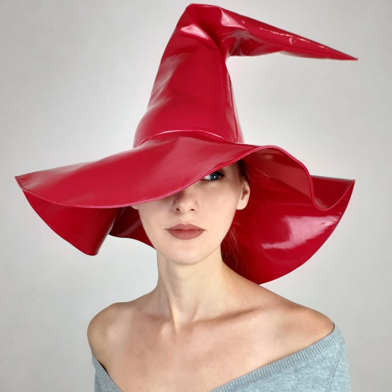 May include: A bright red, glossy, wide-brimmed witch hat with a tall, pointed crown. The hat is worn by a person with long, dark hair.