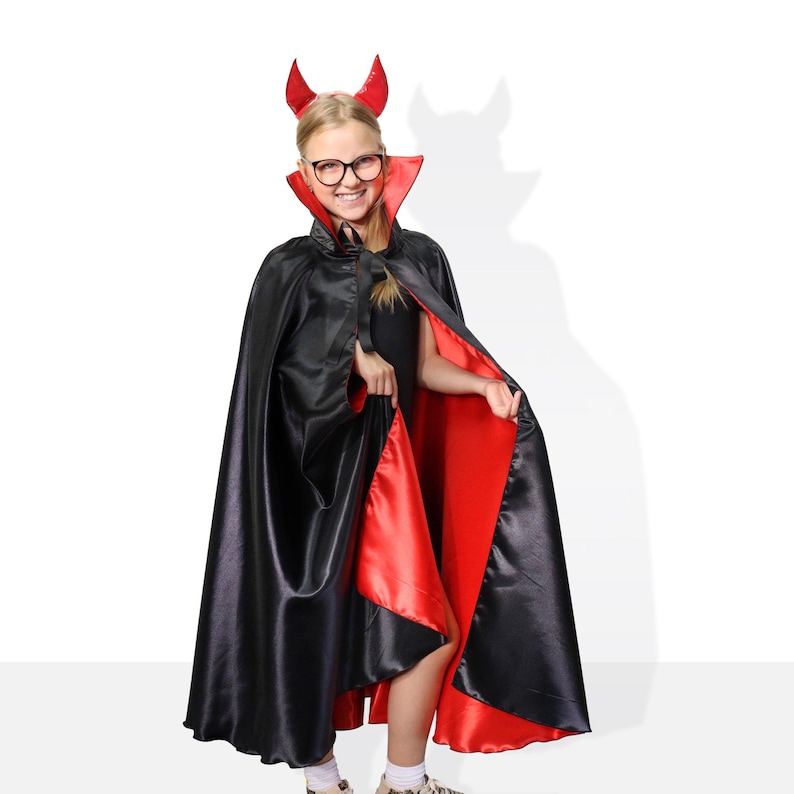 May include: A young person dressed as a devil for Halloween. They are wearing a black cape with a red lining and a red devil horn headband.