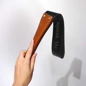 May include: A wooden paddle with a dark brown handle and a black leather strap. The text "Spank Me" is printed on the strap. The paddle is held by a hand against a white background.