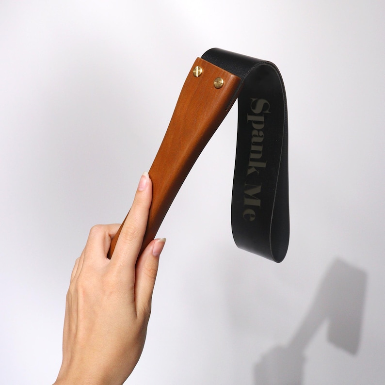 May include: A wooden paddle with a dark brown handle and a black leather strap. The text "Spank Me" is printed on the strap. The paddle is held by a hand against a white background.