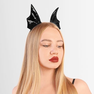 May include: A woman with long blonde hair wearing a black headband with bat wings. She is wearing red lipstick and looking down.