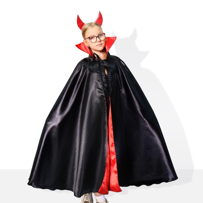 May include: A child dressed as a devil for Halloween. They are wearing a black cape with a red lining and red devil horns. The cape has a red collar. The child is wearing glasses.