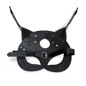 Puede incluir: M&aacute;scara de gato de cuero negro con una correa y una hebilla de metal. La m&aacute;scara tiene un agujero para los ojos y la nariz, y est&aacute; decorada con tachuelas plateadas.