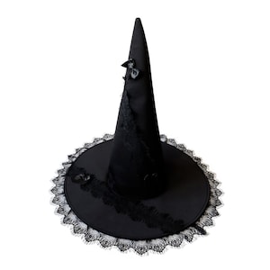 K&ouml;nnte beinhalten: Ein schwarzer Hexenhut mit einer hohen, spitzen Krone und einer breiten Krempe. Die Krempe ist mit schwarzer Spitze besetzt und der Hut ist mit schwarzen Satinb&auml;ndern verziert.
