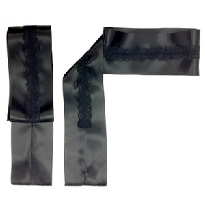 Puede incluir: Cinta de satén negro con delicado ribete de encaje negro. La cinta está doblada y dispuesta para mostrar el detalle del encaje. El satén tiene un ligero brillo y el encaje añade un toque de elegancia. La cinta está sobre un fondo blanco.