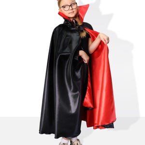 May include: A child wearing a black and red satin cape with a red collar. The cape is long and flowing, and the child is looking to the side.
