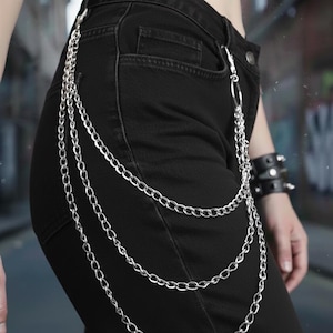 May include: Silver chain accessory with three strands, attached to black denim pants. The chains hang down the side of the pants. The accessory is a stylish addition to any outfit.