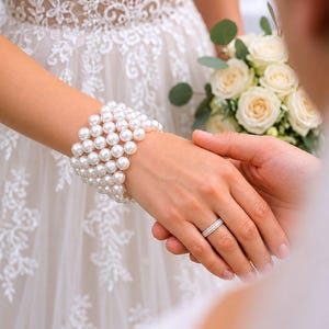May include: A bride's hand adorned with a pearl bracelet and a diamond ring, clasped by another hand. The bride wears a white lace wedding dress and holds a bouquet of white roses. The image captures a moment of a wedding ceremony.