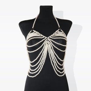 K&ouml;nnte beinhalten: Eine wei&szlig;e Perlen-K&ouml;rperkette mit mehreren Reihen von Perlen, die durch silberne Metallglieder verbunden sind. Die Kette ist so konzipiert, dass sie als Oberteil oder Bralette getragen werden kann.