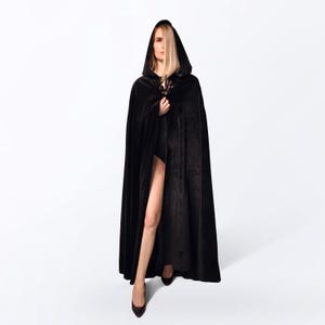 May include: A woman wearing a long black velvet cape with a hood. The cape is open in the front, revealing her legs. She is standing on a white background.