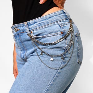 May include: Silver-toned chain belt with multiple strands and heart-shaped charms. The belt is attached to the waistband of light blue denim jeans. The chain has a chunky, industrial design.