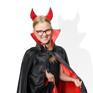 May include: A young person dressed as a devil for Halloween. They are wearing a black cape with a red lining and red devil horns. They are smiling and looking at the camera.
