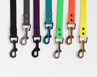 May include: A collection of colorful dog leashes in black, gray, purple, teal, green, orange, and yellow. Each leash has a matching metal clasp and rivets. The leashes are made of a durable, flexible material.