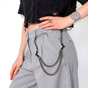 May include: A silver-toned chain wallet accessory with a bat-shaped charm and two parallel chains. The chain is attached to gray pants. The person is wearing a black distressed shirt and a silver watch.