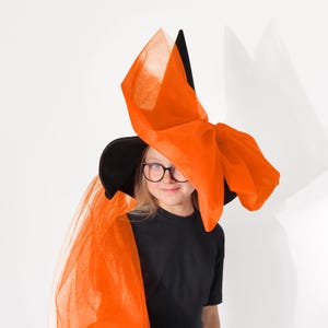 May include: A black witch hat with a large orange tulle bow. The hat is worn by a person with blonde hair and glasses. The tulle bow is tied in a large knot on top of the hat.