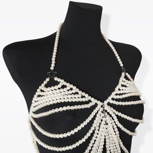 K&ouml;nnte beinhalten: Ein wei&szlig;er Perlenketten-K&ouml;rper-Schmuck mit einem Stern-Anh&auml;nger. Die Kette besteht aus mehreren Perlenstr&auml;ngen und ist so konzipiert, dass sie als Oberteil oder BH getragen werden kann.