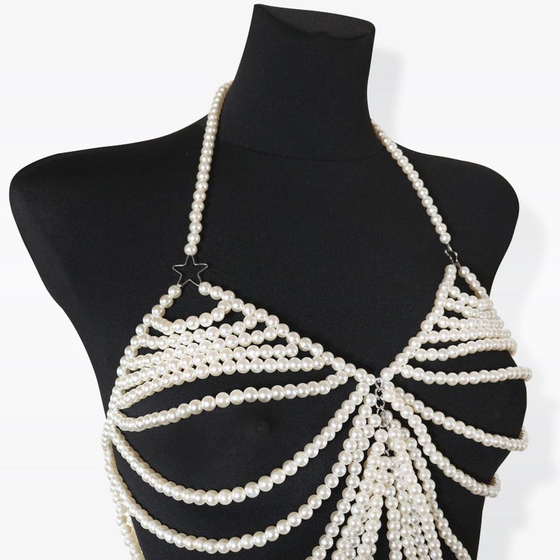 K&ouml;nnte beinhalten: Ein wei&szlig;er Perlenketten-K&ouml;rper-Schmuck mit einem Stern-Anh&auml;nger. Die Kette besteht aus mehreren Perlenstr&auml;ngen und ist so konzipiert, dass sie als Oberteil oder BH getragen werden kann.