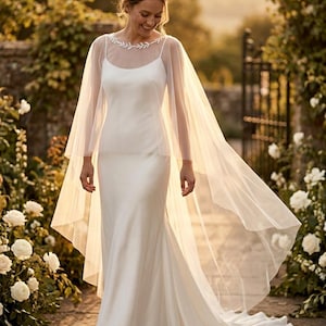 May include: A white wedding dress with a flowing skirt and a sheer, long-sleeved cape. The dress has a simple, elegant design. The bride is walking in a garden setting with white roses and a stone path.