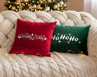 May include: Two square Christmas pillows on a cream-colored sofa. One pillow is red with the text "Holly Jolly" in white script and festive icons. The other is green with "HoHoHo" in white script. A Christmas tree with lights is in the background.