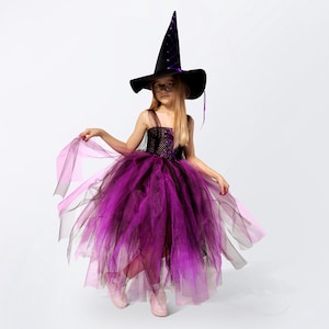 May include: A young girl dressed as a witch for Halloween. She is wearing a purple and black tulle dress with a black witch hat and glasses.