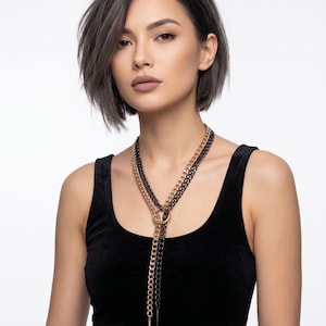 Puede incluir: Un collar de capas con cadenas doradas y negras. El collar tiene un conector circular y un colgante de punta. La modelo lleva una camiseta sin mangas negra. La marca "VERVE CHAINWEAR" es visible en la parte inferior.