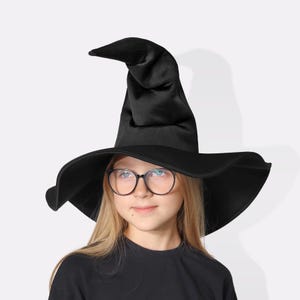 May include: A black witch hat with a wide brim and a pointed top. The hat is made of a soft, possibly satin-like material. The hat is worn by a person wearing a black shirt and glasses.