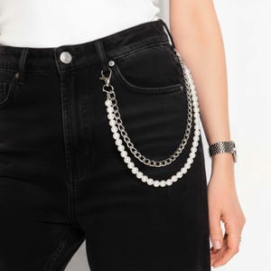 May include: A silver-tone chain with faux pearls and a curb chain design, attached to black denim jeans. The chain is a two-strand design, with a lobster clasp at the top. A silver watch is visible on the wrist.